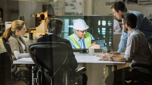 Hybrid work teleconference among several employees in an office and a worker in a hardhat on video.