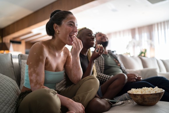 Happy viewers eat popcorn on a couch while watching TV.