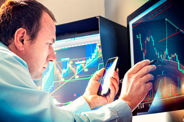 A money manager using a smartphone and stylus to analyze a stock chart on a computer monitor.