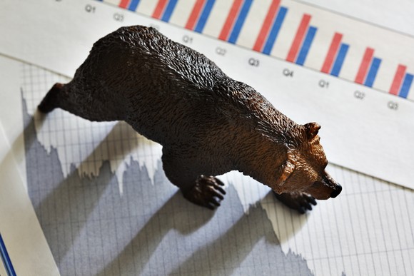 A bear figurine walking across a newspaper clipping of a plunging chart and quarterly financial metrics.