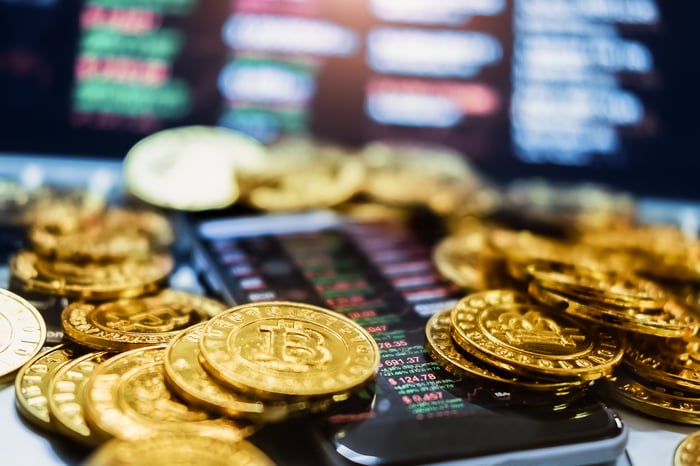 Gold coins stamped with the Bitcoin logo set atop a smartphone displaying crypto quotes and charts. 