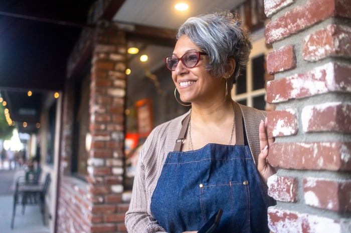 A smiling person in an apron standing outside a brick building.