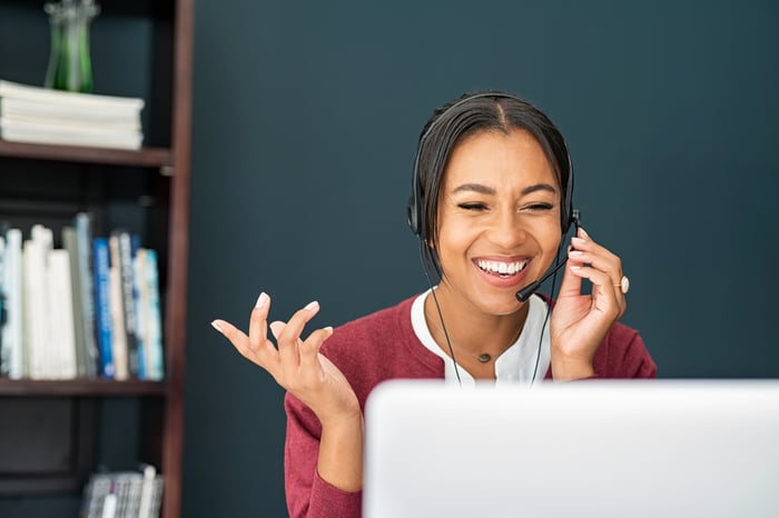 A smiling person speaking on a telephone headset while looking at a laptop. 