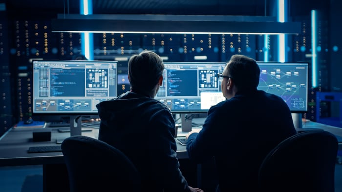 Two employees looking at copious amounts of data on three computer monitors in a dark room. 