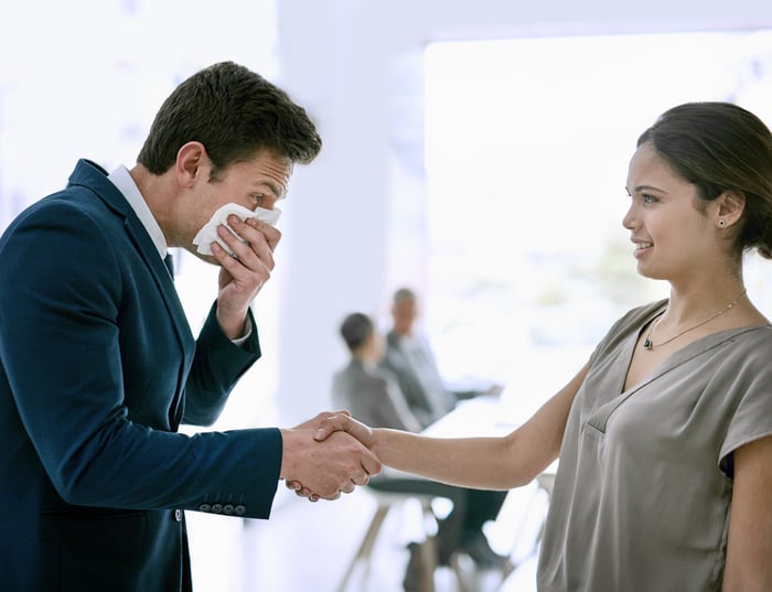 A business person holds a tissue to their face while shaking hands with another person.