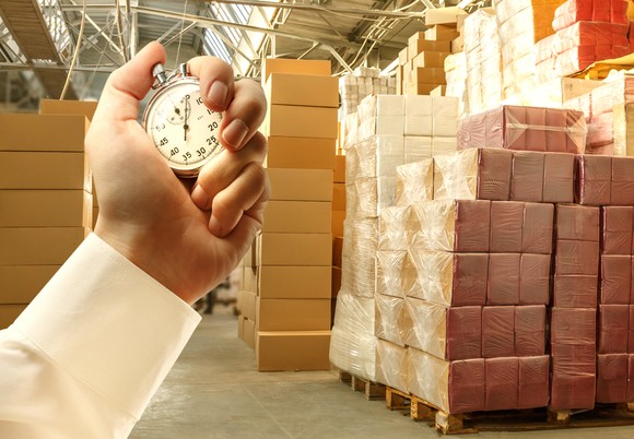A sleeved hand holding a stopwatch in front of stacks of boxes in a warehouse.