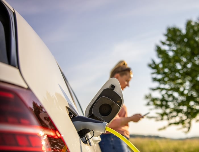 Woman standing near her white EV as it charges in a rural environment.