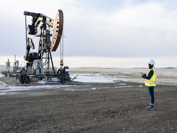 An oilfield worker wearing personal protective equipment holds a clipboard next to a pumpjack.