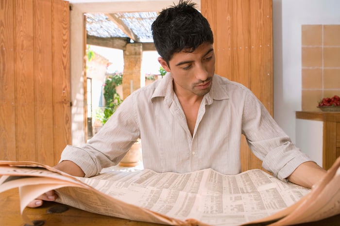 A person reading a financial newspaper while seated at a table.