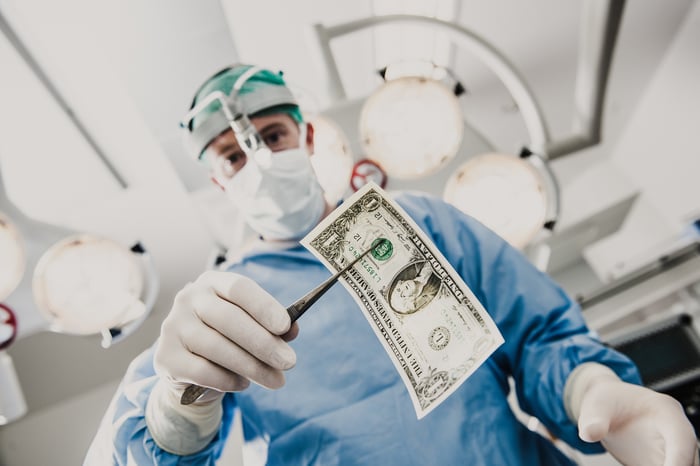 A surgeon in an operating room using forceps to hold a $1 bill.