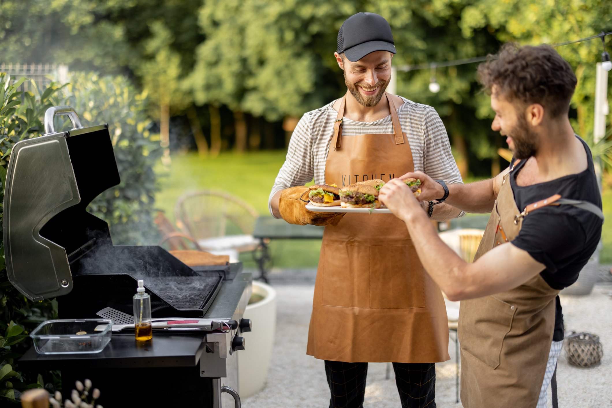 two people smile in an outdoor setting while eating recently grilled hamburgers