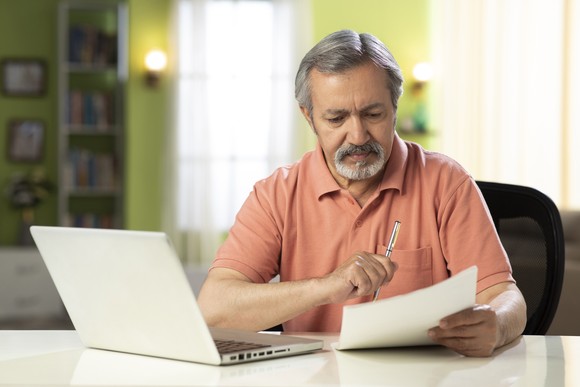 A person at a table with an open laptop holding a pen and document.