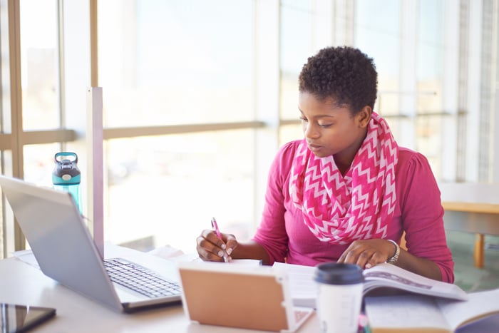 Someone reading a textbook and writing in front of an open laptop.