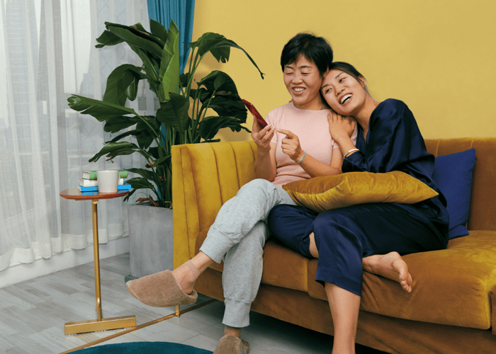 Two smiling people sitting on a couch while one looks at their smartphone.