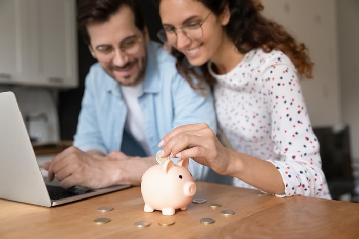 A man and a woman sit next to each other. The woman inserts coins into a piggy bank.