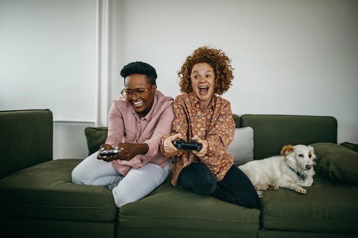 Two people play a video game while sitting on a couch next to a dog.