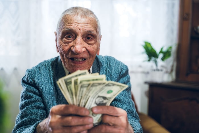 A smiling person who's sitting in a chair and holding up a fanned assortment of cash bills. 