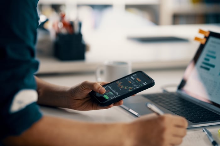 Close-up of person looking at stock chart on cellphone while writing on piece of paper.