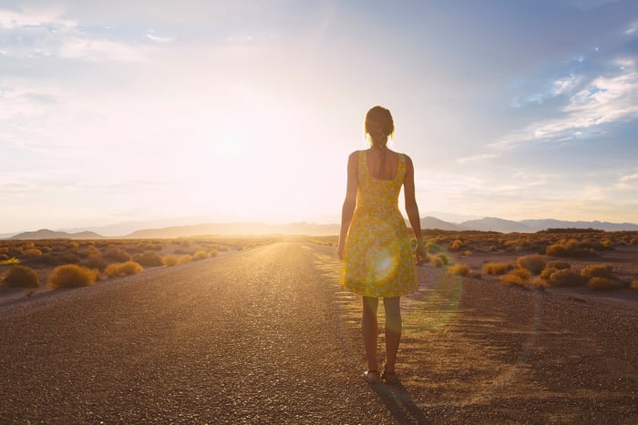 A person stands on an open road with their back to the viewer while looking at the rising sun. 