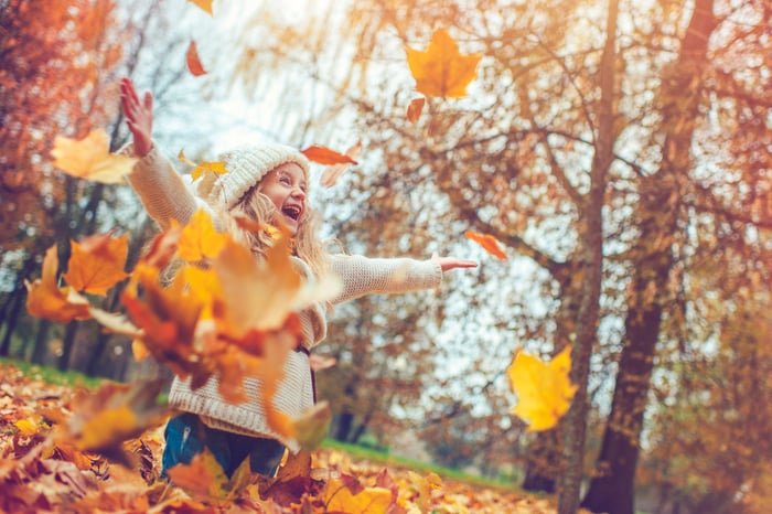 A child smiles while playing in the red, orange, and yellow autumn leaves. 