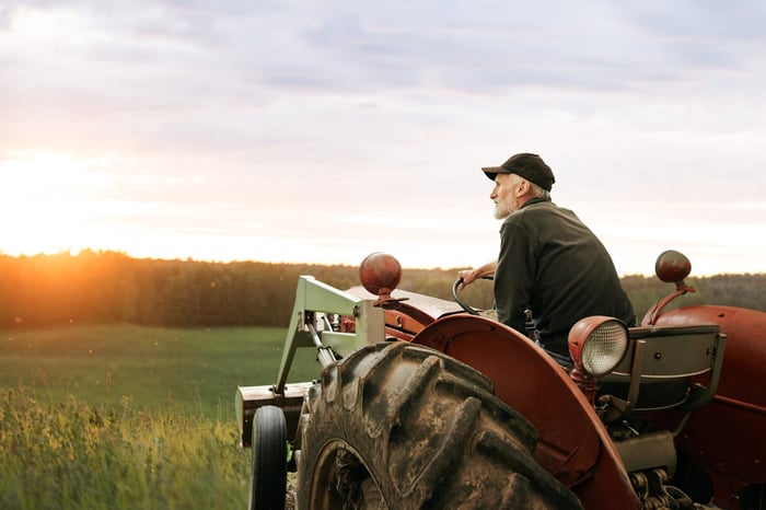 A person sitting on a tractor looks out into an open field with a setting sun over the tree line.