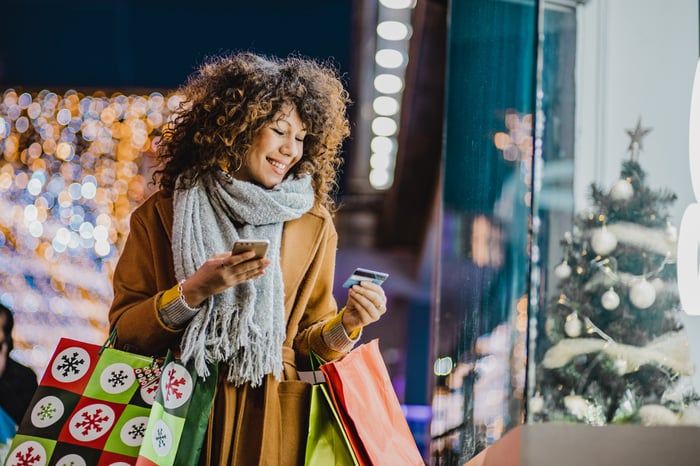 A person carrying Christmas presents while looking at a smartphone and credit card.