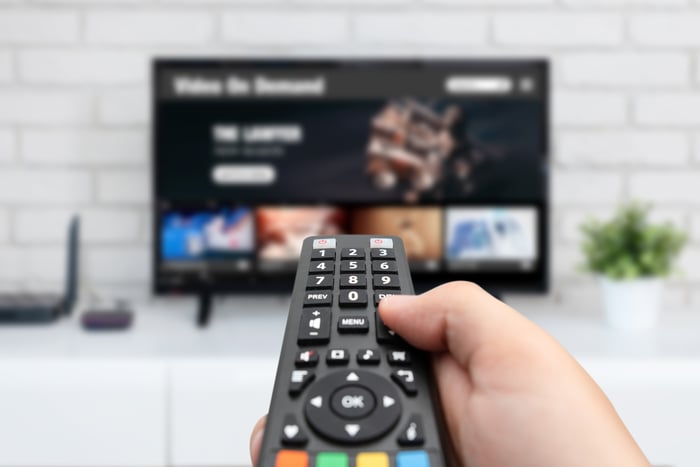 A hand holds a remote in front of a TV displaying streaming options.