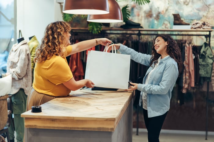 A happy store clerk hands a shopping bag to a smiling customer. 