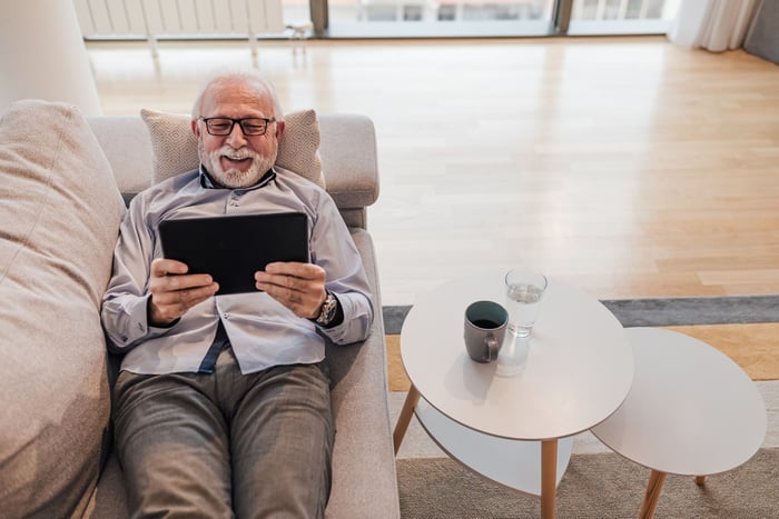 A smiling person lying on a couch holding a tablet.