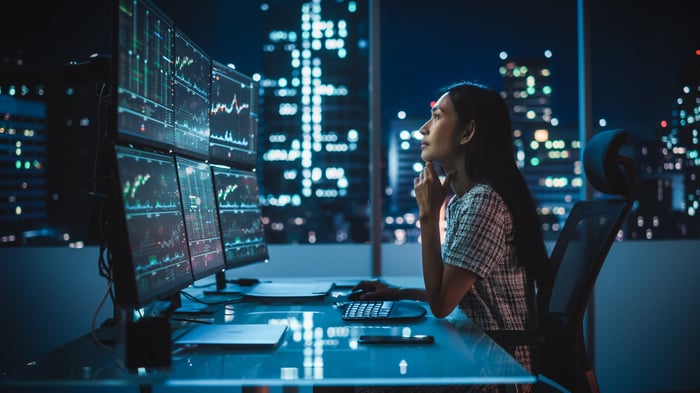 A person looks at six monitors showing stock charts with a darkened city skyline in the background.