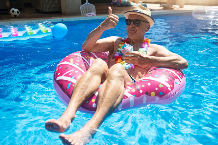 Person giving the thumbs-up sign and holding a drink while floating in an inner tube in a pool.