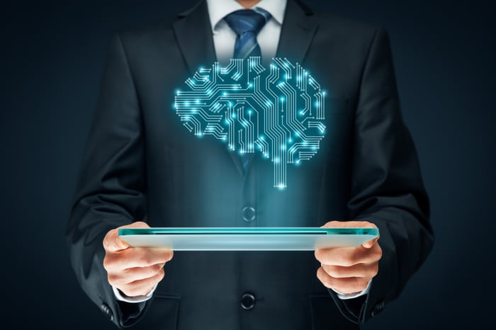 A hologram of a digital brain with visible pathways above a tablet that's being held by a businessperson.