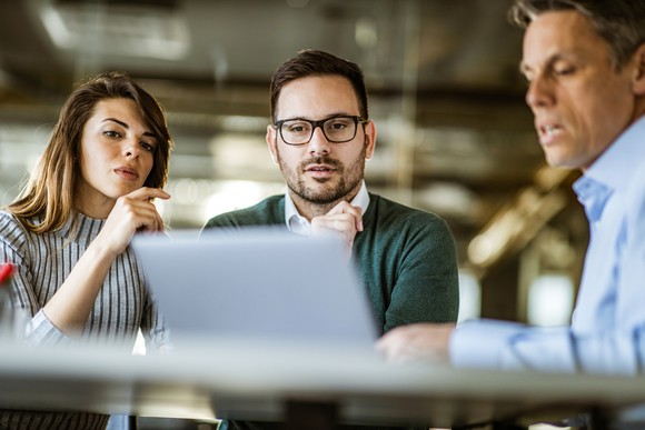 Three investors gather around a laptop while one points at the screen and makes a comment.
