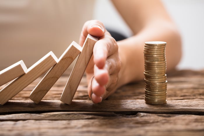 A hand stopping falling dominos from overturning a stack of coins.