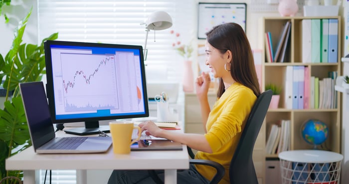 A smiling person looking at a monitor showing a stock chart trending upward.