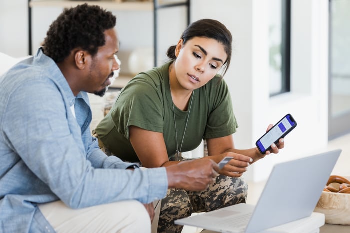 Two people in a living room look at a phone and computer together.