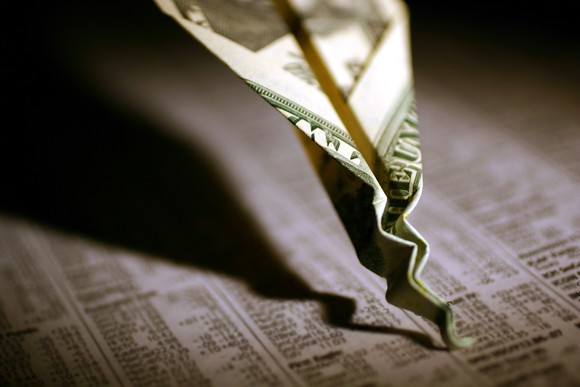 A twenty dollar bill paper airplane that's crashed and crumpled into a financial newspaper with visible stock quotes.