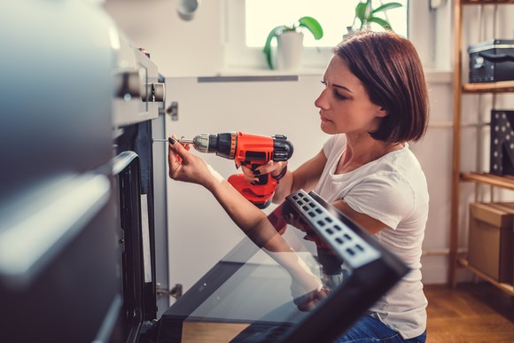 A person uses a power tool to fix an appliance in a room with a window in the background.
