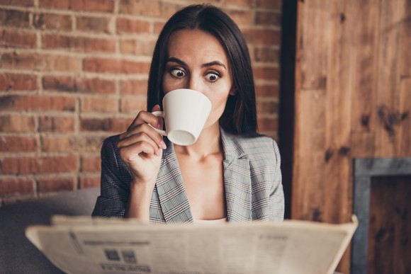 Person reading a newspaper and drinking from a cup.