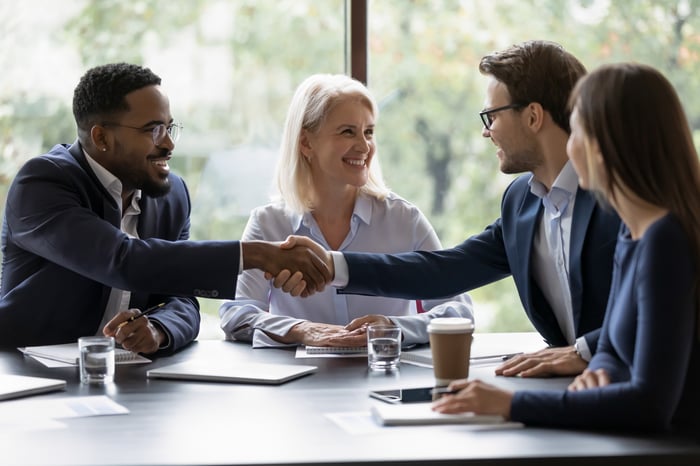 Professionals in a business environment shake hands at a conference table. 