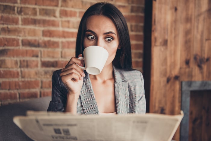 Astonished woman looks at newspaper while drinking coffee