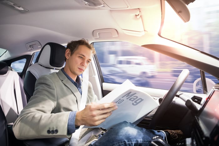 A businessperson reading a magazine while behind the wheel of an autonomous vehicle. 