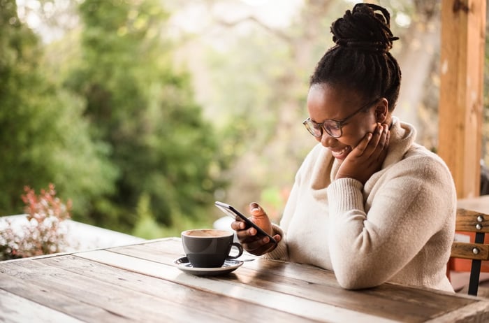 A person smiles while looking at their phone and enjoying a hot beverage while sitting at a table.