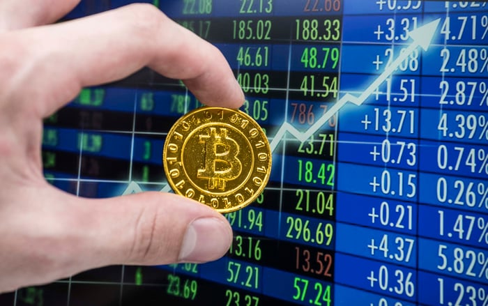 A hand holding a gold token with the Bitcoin logo on it in front of a series of stock movements.