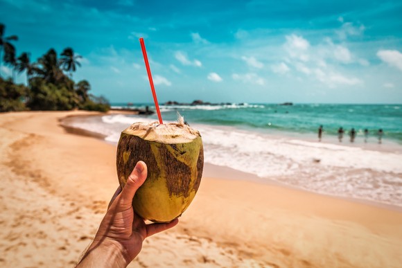 A person on a beach holding up a sliced coconut with a straw in it.