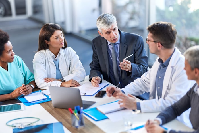 A group of doctors talk with a coworker while sitting at a conference table.