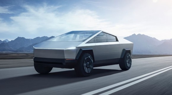 A Tesla Cybertruck driving down a single-land road, with mountains in the background.