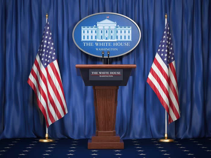 American flags flank a podium in front of a blue curtain and a White House logo.