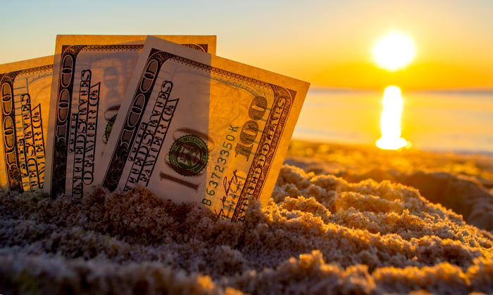 Three one hundred dollar bills partially buried in sand, with the sun blazing over the ocean in the background.