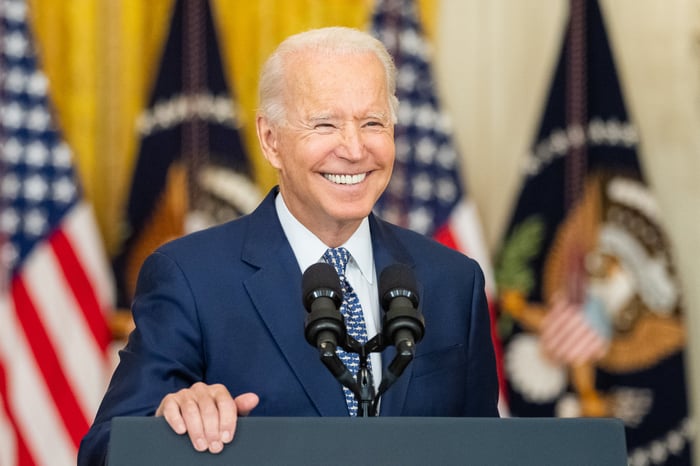 A smiling President Biden standing behind the presidential podium in the East Room of the White House.